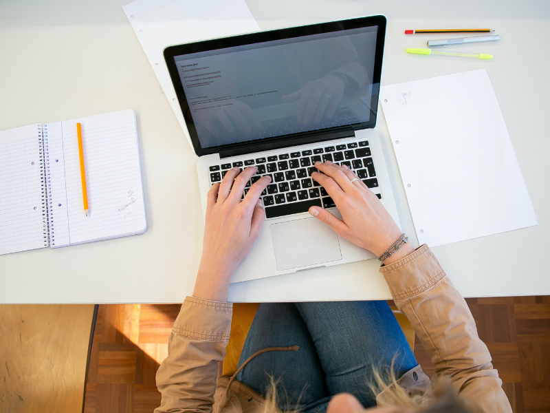 A view of arms and hands typing on a laptop from above, with notes on the table beside the laptop.